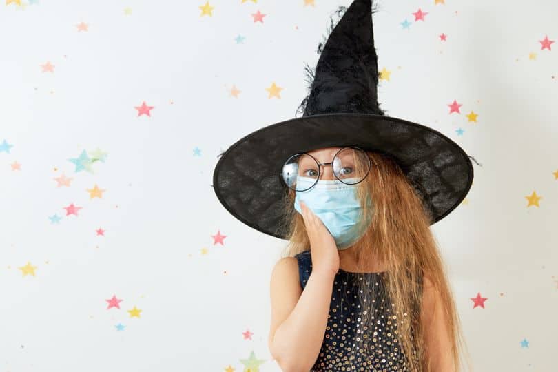 A young girl dressed in a witch costume and a surgical mask.